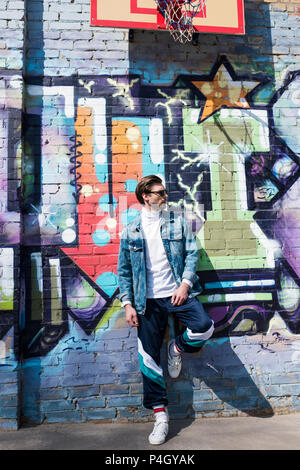 Beau jeune homme en vêtements vintage se penchant sur mur de brique avec graffiti Banque D'Images