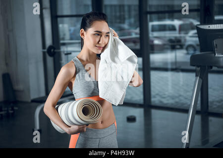 Jeune femme sportive avec tapis de yoga roulé la sueur avec une serviette après essuyage des exercices dans la salle de sport Banque D'Images