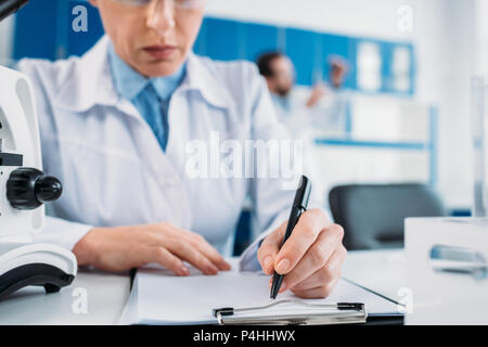 Vue partielle de female scientist prendre des notes dans le bloc-notes sur le lieu de travail avec collègue derrière in lab Banque D'Images