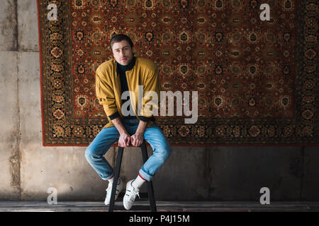 Jeune homme en veste jaune assis sur une chaise en face de pendaison tapis sur le mur Banque D'Images