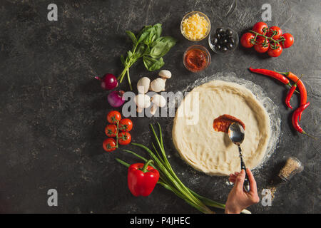 Cropped shot of woman putting sauce sur pâte crue pendant la cuisson de la pizza italienne sur la table sombre Banque D'Images