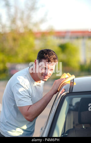 Portrait of a smiling young man cleaning sa voiture avec un chiffon en microfibre à l'extérieur Banque D'Images