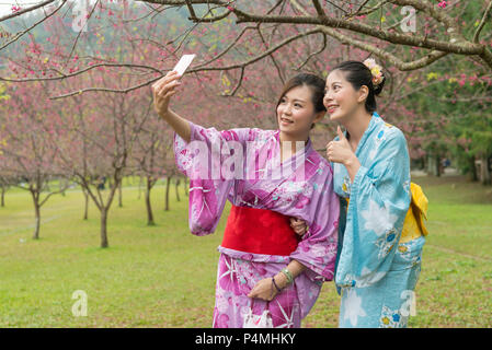 Deux jeunes femmes asiatiques en tenant à l'aide de leur téléphone cellulaire selfies en vertu de la cherry blossom tree. Une holding cell phone une autre posant son pouce. Banque D'Images