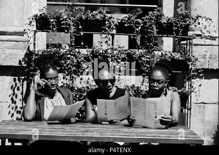 Élégant trois african american womans posés au jour d'été ensoleillé extérieur, sitting on table de restaurant avec menu à la main. Banque D'Images