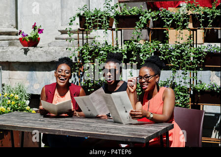 Élégant trois african american womans posés au jour d'été ensoleillé extérieur, sitting on table de restaurant avec menu à la main. Banque D'Images