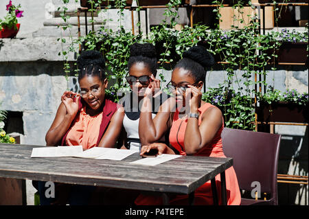 Élégant trois african american womans posés au jour d'été ensoleillé extérieur, sitting on table de restaurant avec menu à la main. Banque D'Images