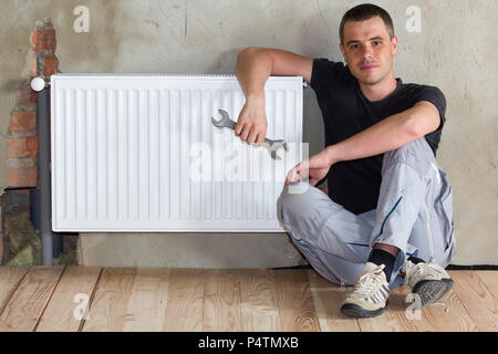 Jeune beau plombier assis sur marbre avec une clé dans la main près du radiateur de chauffage installé avec succès en salle vide d'un appartement nouvellement construit ou h Banque D'Images