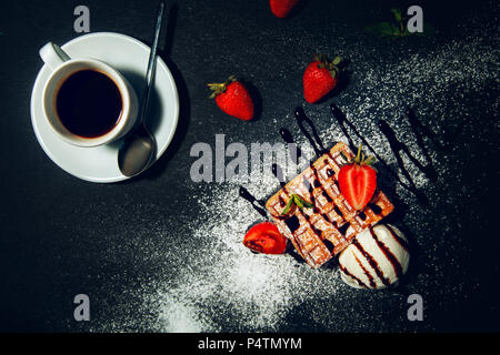 Tasse de café avec des gaufres belges avec de la crème glacée et les fraises sur un fond noir. Banque D'Images