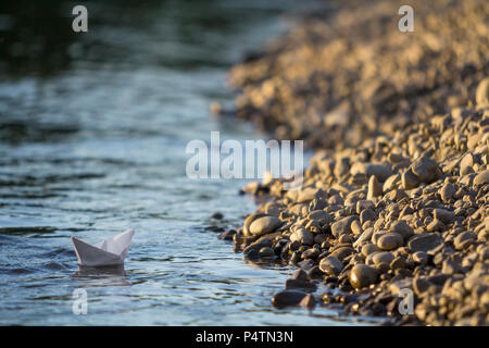 De simples petits white paper toy origami bateau flottant sur la rivière claire ou l'eau de mer sur l'eau bleu et marron floue fond de plage de galets. La beauté de nat Banque D'Images