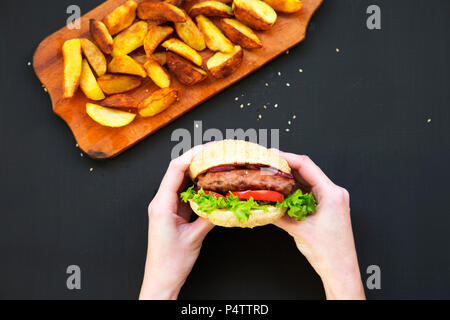 Jeune fille est titulaire d'hamburger. Pommes de terre rissolées sur planche de bois. Vue d'en haut. Banque D'Images