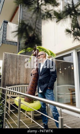 Senior businessman debout dans son jardin, tenant une palme en caoutchouc, rêvant de vacances Banque D'Images