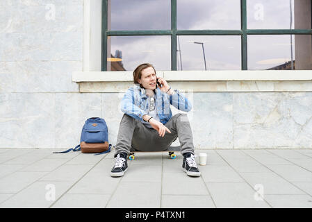Jeune homme avec skateboard assis sur la masse, using smartphone Banque D'Images