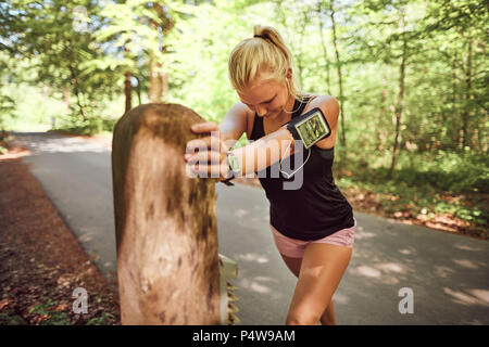 Fit young blonde woman in sportswear appuyée sur un poster à côté d'un chemin forestier qui s'étend avant d'aller pour une course Banque D'Images