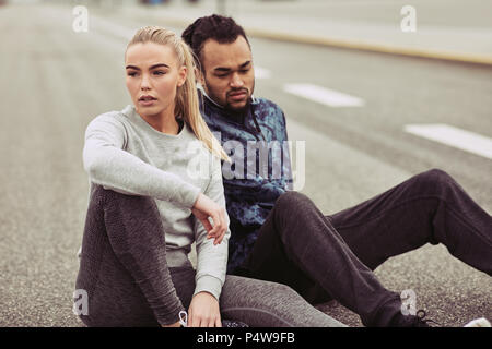 Fatigué à la young couple in sportswear assis sur une route reprendre leur souffle tandis que sortir courir ensemble Banque D'Images
