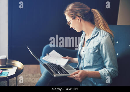 Smiling young businesswoman reading plus de documents de travail en ligne avec un ordinateur portable tout en étant assis sur un canapé Banque D'Images