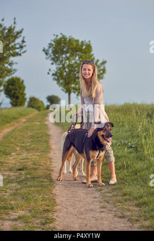 Jolie femme blonde souriant avec ses deux chiens sur conduit debout sur un chemin de terre dans les terres agricoles à la recherche de la caméra dans la lumière du soir Banque D'Images