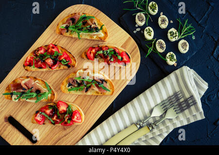 Bruschetta italienne avec tomates, jambon de Parme, roquette et sauce balsamique glasse sur une planche à découper et le fromage mini bouchées sur dark backgroun Banque D'Images