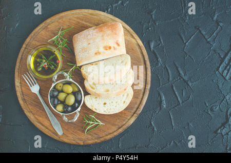 En-cas méditerranéens. Olives vertes et noires, d'huile d'olive, d'herbes et de tranches de pain ciabatta sur planche de bois sombre sur fond de pierre. La cuisine Italienne Banque D'Images