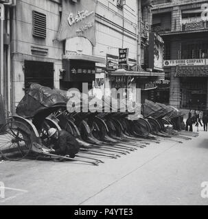 Années 1950, historique, garée dans une petite rue de la vieille ville de Hong Kong, une ligne de rickshaws avec des capots.Ces chariots de transport de passagers à deux roues motorisés par un homme, où une personne tire une autre personne, étaient une forme de transport populaire dans de nombreuses villes asiatiques.Le mot rickshaw est originaire du mot japonais pour « véhicule à moteur humain ».Le pousse-pousse original est maintenant connu comme un pousse-pousse tiré, car il a été remplacé dans la plupart des cities asiatiques par d'autres formes de pousse-pousse, notamment le pousse-pousse à cycle et plus récemment le pousse-pousse électrique. Banque D'Images