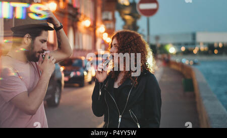 Couple aimant de hipsters sur date. Guy en chapeau et t-shirt fume cigarette électronique. Fille aux cheveux roux bouclés est holding cigarette electronique. Banque D'Images