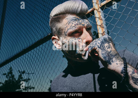 Portrait de jeune homme tatoué fumer une cigarette à Wire Mesh fence Banque D'Images