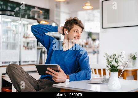 L'homme souriant dans un café avec écouteurs, ordinateur portable et tablette Banque D'Images