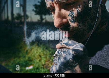 Portrait de jeune homme tatoué avec écouteurs fumer une cigarette Banque D'Images