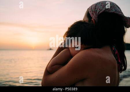 Petite fille mère tenant sur ses bras en face de la mer, regardant le coucher du soleil Banque D'Images