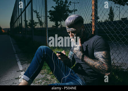 Jeune homme tatoué avec écouteurs et smartphone fumer une cigarette à Wire Mesh fence Banque D'Images