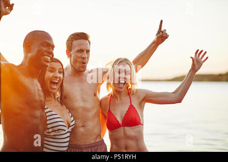 Groupe de rire insouciant young friends standing in arm in arm ensemble tout en s'amusant au bord d'un lac en fin d'après-midi Banque D'Images