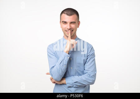 Jeune homme latin en chemise bleue garde doigt sur l'avancement des lèvres, tente de garder la conspiration. Essayez de garder secrets. Capture d isolé homme séduisant sur zone de gris Banque D'Images