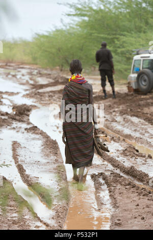 Kakuma, Kenya Turkana - une femme marche pieds nus sur un chemin de terre humide avec la pluie. Dans l'arrière-plan un un Land Rover coincé dans la boue. Banque D'Images