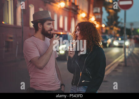 Couple aimant de hipsters sur date. Guy en chapeau et T-shirt est holding electronic cigarette. Fille aux cheveux roux bouclés organise une e-cigarette. Ils ar Banque D'Images