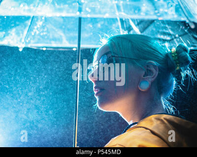 Très jolie fille aux cheveux teints en bleu imperméable jaune et transparent avec des socles de parasol près de la fontaine. Néon nuit de ville. Portrait de hipster élégant avec des lunettes. Banque D'Images