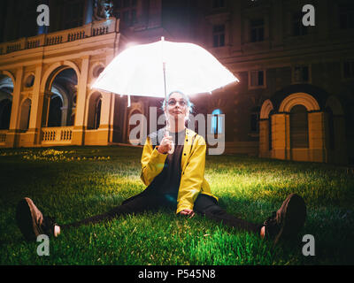 Très jolie fille aux cheveux teints en bleu imperméable jaune et avec parapluie transparent et lumineux assis sur l'herbe verte près de l'édifice historique. Portrait de hipster élégant avec des lunettes. Banque D'Images