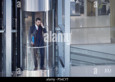 Businessman dans l'ascenseur en verre moderne de parler par le téléphone et la tenue de documents Banque D'Images