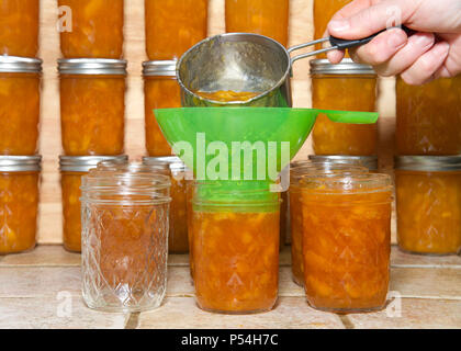 La femme plus âgée avec godet de mesure pleine de confiture de pêches fraîchement préparés de passer par l'entonnoir vert en bocaux de conserve avec pots empilés en rangées fini Banque D'Images