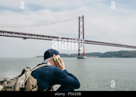 Un jeune voyageur ou touriste avec un sac à dos sur le bord de l'eau à Lisbonne au Portugal, à côté de la pont du 25 avril. Il parle au téléphone Banque D'Images