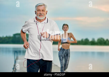 Senior man listening music, tournant près du lac en soirée. Jeune fille courir derrière. Activités de plein air, mode de vie sain, fort compétents, mettre en place des chiffres. Différentes générations. Le sport, yoga, fitness Banque D'Images