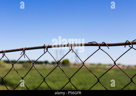 Old rusty wire mesh fence contre l'arrière-plan flou d'un champ vert, un silos à grains et un ciel bleu clair Banque D'Images