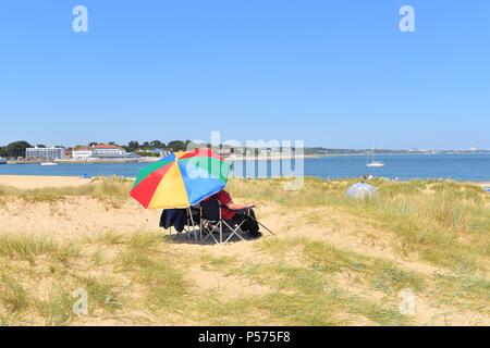 Couple assis dans des chaises longues sous un parasol à rayures colorées au cours de 2018 sur la vague de south Dorset Coast Beach de Studland, UK Banque D'Images