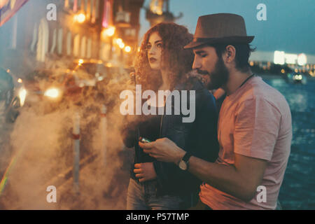 Couple aimant de hipsters sur date. Guy en chapeau et T-shirt est serrant sa petite amie. Fille aux cheveux roux bouclés fume cigarette électronique. Banque D'Images