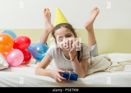 L'anniversaire d'une adolescente est de 10-11 ans. Une fille dans un chapeau de fête avec un cadeau se trouve sur le lit dans une chambre d'enfants, des ballons d'arrière-plan Banque D'Images