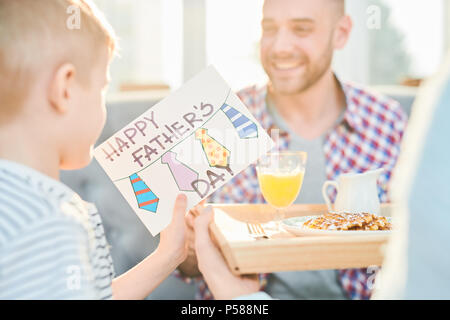 Tourné aux teintes chaleureuses du petit garçon à la main de son père donnant des pères et de la surprise de la carte petit-déjeuner à home, copy space Banque D'Images