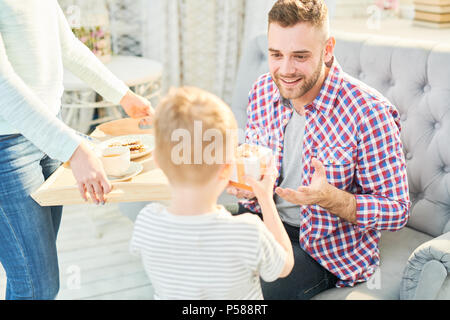 Portrait aux teintes chaleureuses du beau jeune homme à la tendresse à son fils lui donnant le présent et petit-déjeuner surprise pour la fête des pères à la maison, copy space Banque D'Images