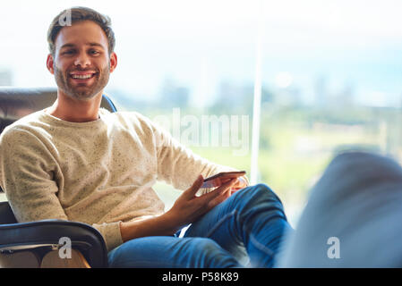 Portrait homme assis sur un canapé moderne à côté de grandes fenêtres en verre avec une vue imprenable derrière lui comme il sourit à l'appareil photo avec son téléphone toujours détenus dans ses mains. Banque D'Images