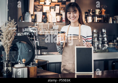 Asian female barista porter jean apron holding Coffee cup et blank blackboard menu café au comptoir bar avec smile face,cafe service concept,business Banque D'Images