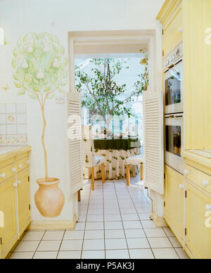 Arbre en pot au pochoir sur le mur de cuisine avec unités jaune pâle et vue de salle à manger par porte ouverte Banque D'Images