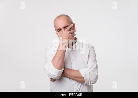 Déplut homme mûr en chemise blanche couvrant son visage avec Main sur fond gris. Banque D'Images