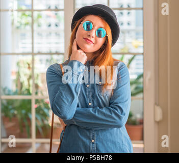 Rousse élégante femme portant chapeau melon et des lunettes with hand on chin la réflexion sur la question, l'expression pensive. Smiling avec visage. D Banque D'Images